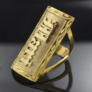 May include: A gold-colored ring with a rectangular face. The face features the word "HIS" in a stylized font, set against a textured background. The ring has a decorative border and a polished finish.
