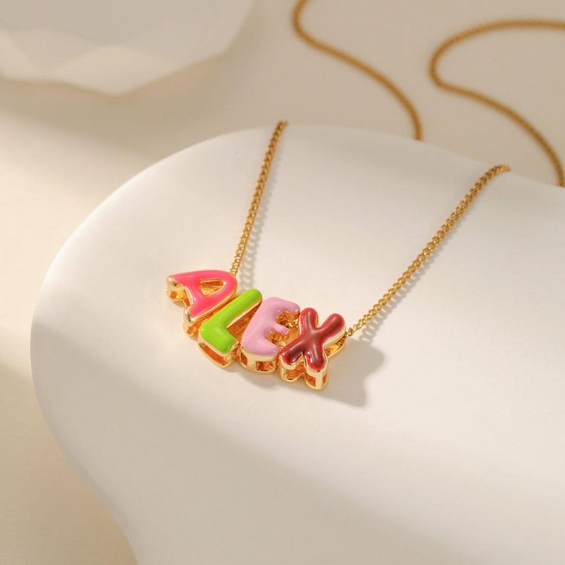 May include: A gold-colored necklace with the name "ALEX" spelled out in colorful, block letters. The letters are pink, green, light pink, and maroon. The necklace is on a white surface.
