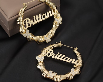 Pode incluir: Brincos de argola dourados com o nome "Brittany" escrito em itálico e detalhes de borboleta com strass transparentes.