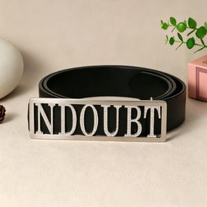 May include: A black leather belt with a silver buckle that spells out the word "NDOUBT" in capital letters. The letters are filled with small, clear rhinestones. The belt is displayed on a neutral surface.