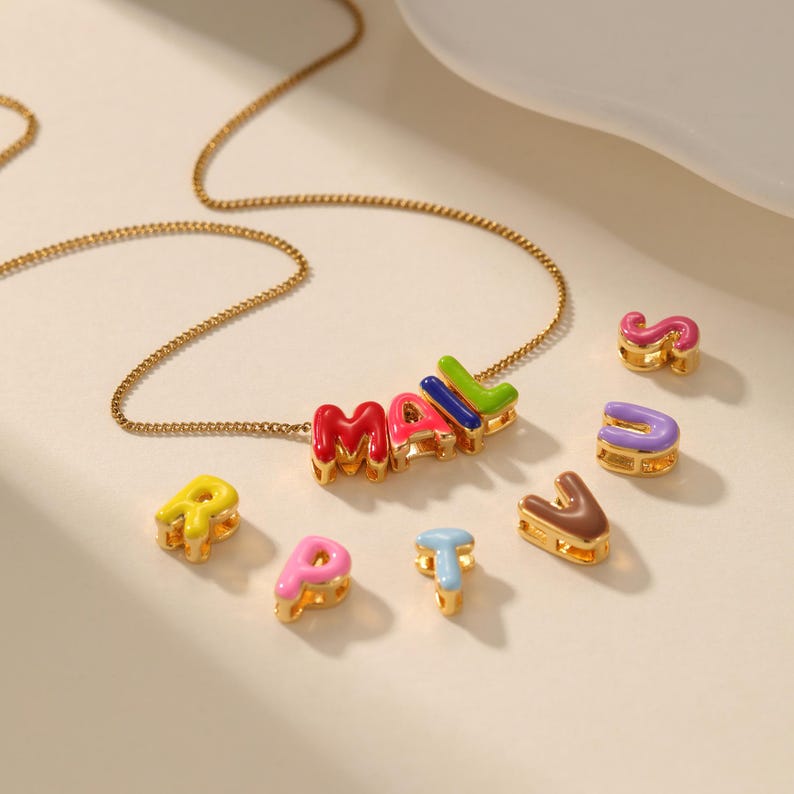 May include: A gold-colored necklace with the name "MALL" spelled out in colorful enamel letters. Additional individual letters in various colors are scattered around the necklace, including "R", "P", "T", "S", "U", and "V".