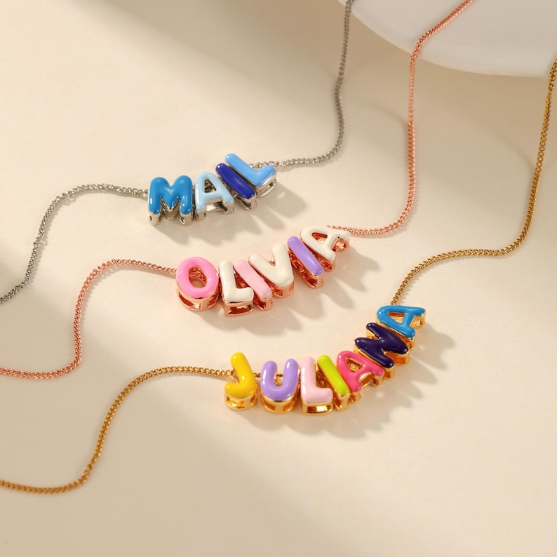 May include: Three colorful name necklaces. The necklaces feature the names "MAX", "OLIVIA", and "JULIANA" spelled out in vibrant, block-style letters. Each name is a different color, with a matching chain. The necklaces are displayed on a light-colored surface.