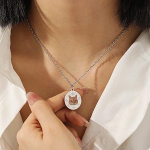 Puede incluir: Collar plateado con un colgante con la cara de un gato. El colgante ovalado presenta una imagen detallada de la cara de un gato atigrado naranja, rodeada de detalles en forma de estrella. El collar se lleva sobre una camisa blanca.