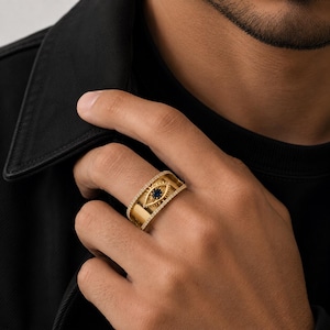 Pode incluir: Um anel de banda larga em ouro com um motivo de olho central e uma pedra azul-escura no meio. As bordas da banda são contornadas por pequenas pedras claras cravadas em pavé, criando uma aparência detalhada e estruturada no dedo.