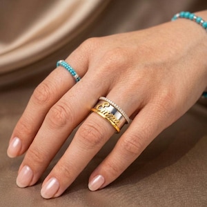 May include: A stack of three rings on a finger, including a thin band with small clear stones, a wide silver-toned band featuring the name Emma in gold-toned script, and a plain gold-toned band. A turquoise beaded ring is on another finger.