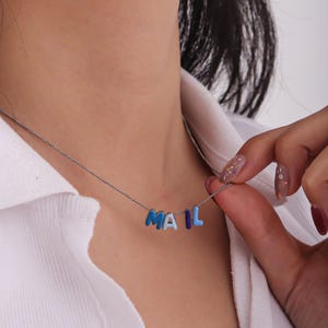 May include: A silver chain necklace with the word "MAIL" spelled out in blue enamel letters. The letters are in varying shades of blue, from light to dark. The necklace is worn against a white shirt.