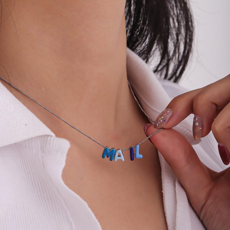 May include: A silver chain necklace with the word "MAIL" spelled out in blue enamel letters. The letters are in varying shades of blue, from light to dark. The necklace is worn against a white shirt.