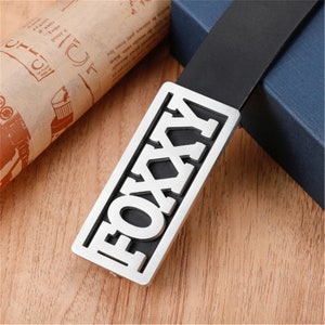 Peut inclure: Une boucle de ceinture argentée avec le mot "FOXXY" découpé sur un fond noir.