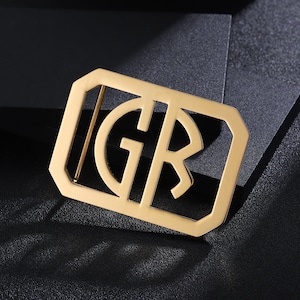 Puede incluir: Una hebilla de cinturón de metal dorado con las iniciales "GR" recortadas en el centro.