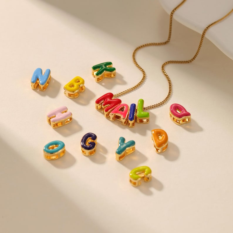 May include: A gold-colored necklace with the word "MAIL" spelled out in colorful enamel letters. Additional individual letters in various colors are scattered around the necklace. The letters are in blue, yellow, green, pink, and orange.