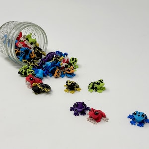 May include: A clear glass jar tipped over, spilling out a collection of colorful frog figurines. The frogs are in various colors, including red, blue, green, yellow, and black. Some have patterned designs. The frogs are scattered on a white surface.