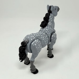 May include: A gray toy horse with white spots, a black mane and tail, and black hooves. The horse is posed in a standing position, with its head up and tail slightly curved. The toy is made of plastic and has articulated joints.