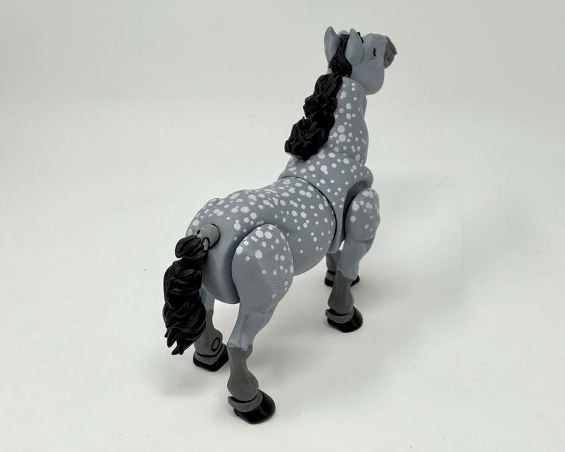 May include: A gray toy horse with white spots, a black mane and tail, and black hooves. The horse is posed in a standing position, with its head up and tail slightly curved. The toy is made of plastic and has articulated joints.