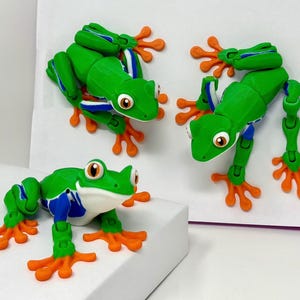 May include: Three green frog figurines with orange feet and large, round eyes. The frogs have articulated limbs and are posed in different positions. The frogs have blue and white accents on their bodies. The background is white.