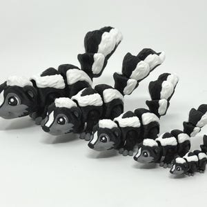 May include: A collection of black and white skunk figurines of varying sizes. Each skunk has a black body with white stripes and a fluffy white tail. The skunks have gray faces and are arranged on a white surface.