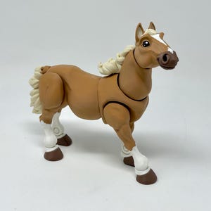 May include: A tan-colored toy horse with a white blaze on its face and white mane and tail. The toy horse has articulated joints and brown hooves. The horse is standing on a white surface.