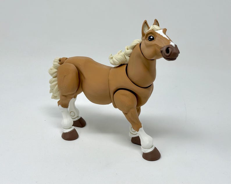 May include: A tan-colored toy horse with a white blaze on its face and white mane and tail. The toy horse has articulated joints and brown hooves. The horse is standing on a white surface.