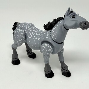 May include: A gray toy horse with white spots and a black mane and tail. The horse has articulated joints and black hooves. The toy is standing on a white surface.