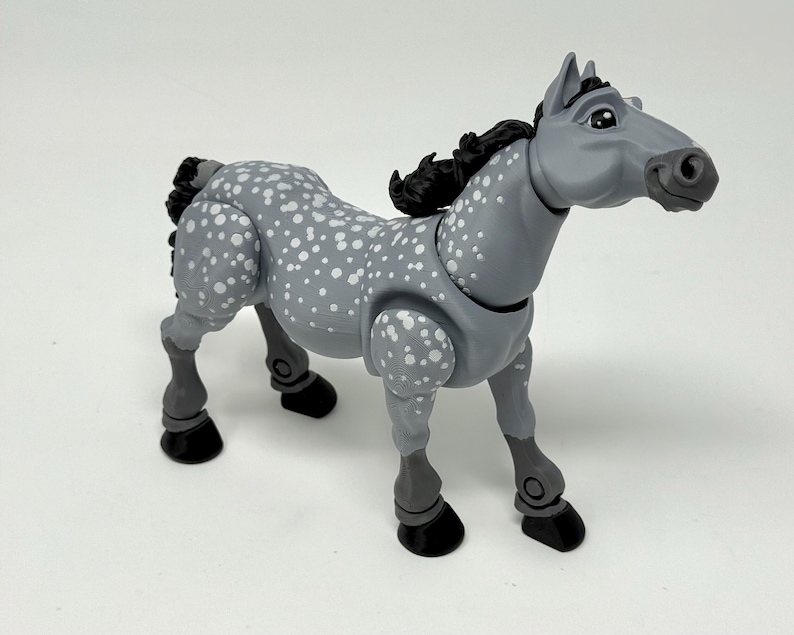 May include: A gray toy horse with white spots and a black mane and tail. The horse has articulated joints and black hooves. The toy is standing on a white surface.
