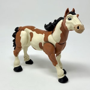 May include: A toy horse figurine with a brown and white spotted coat, black mane and tail, and black hooves. The horse has a light tan muzzle and is posed in a standing position. The toy is made of plastic.