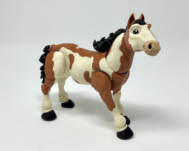 May include: A toy horse figurine with a brown and white spotted coat, black mane and tail, and black hooves. The horse has a light tan muzzle and is posed in a standing position. The toy is made of plastic.