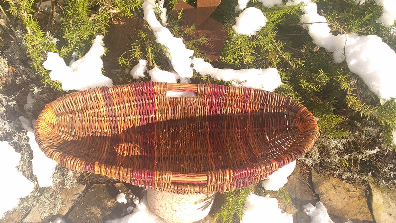 May include: A large, oval-shaped wicker basket with a reddish-brown stain. The basket has two handles and is woven with a tight, intricate pattern.