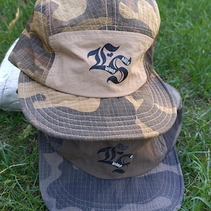 May include: Two five-panel caps stacked vertically. The top cap features a camouflage pattern with a tan front panel displaying the black text "living kills". The bottom cap is a darker grey camouflage with a matching grey front panel and logo.