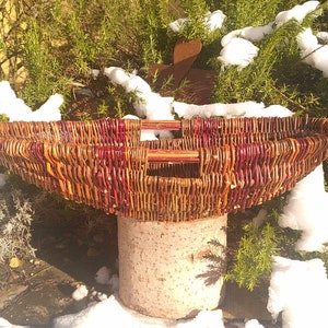 May include: A large, oval-shaped wicker basket with two wooden handles. The basket is woven with brown and red wicker and is sitting on a stone pedestal. The basket is surrounded by snow-covered greenery.