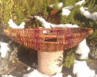 May include: A large, oval-shaped wicker basket with two wooden handles. The basket is woven with brown and red wicker and is sitting on a stone pedestal. The basket is surrounded by snow-covered greenery.