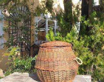 May include: A woven wicker basket with a lid and a rope handle sits on a weathered wooden table with blue paint. The basket is brown and has a natural woven pattern.