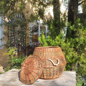 May include: A woven wicker basket with a lid, sitting on a rustic wooden table with chipped blue paint. The basket is made of natural brown wicker and has a rope handle.