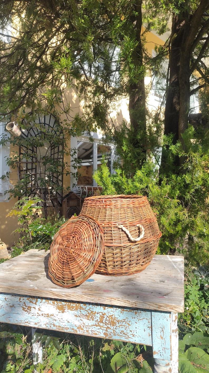 May include: A woven wicker basket with a lid, sitting on a rustic wooden table with chipped blue paint. The basket is made of natural brown wicker and has a rope handle.