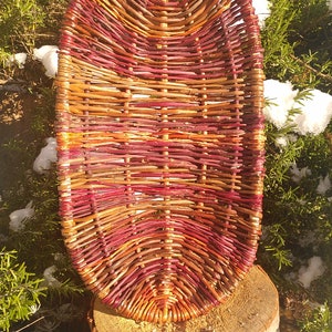 May include: A woven basket made of natural twigs, with a reddish-brown and orange color scheme. The basket is oval shaped and has a smooth, natural finish.