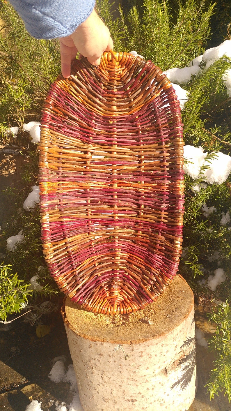 May include: A woven basket made of natural twigs, with a reddish-brown and orange color scheme. The basket is oval shaped and has a smooth, natural finish.