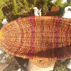 May include: A woven basket with a reddish-brown and brown color scheme. The basket is oval shaped and has a wide, flat base.