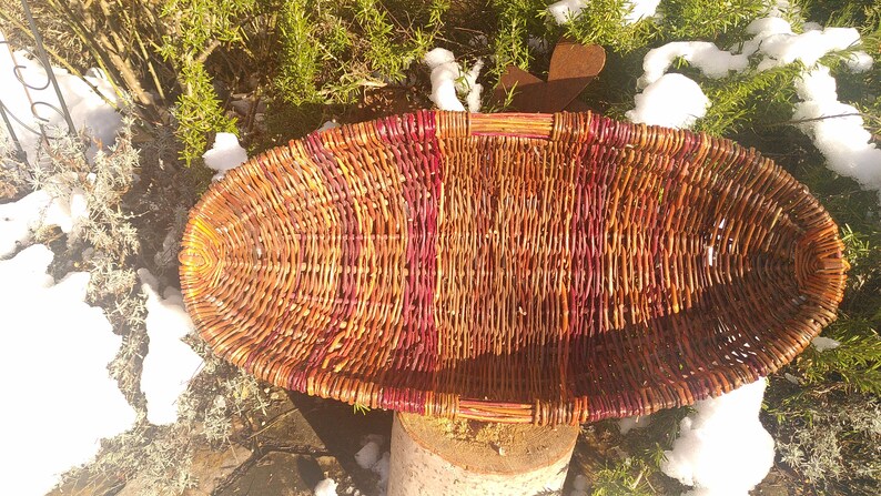 May include: A woven basket with a reddish-brown and brown color scheme. The basket is oval shaped and has a wide, flat base.