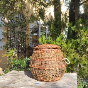 May include: A woven wicker basket with a lid and a rope handle sits on a weathered wooden table with blue paint. The basket is brown and has a natural woven pattern.
