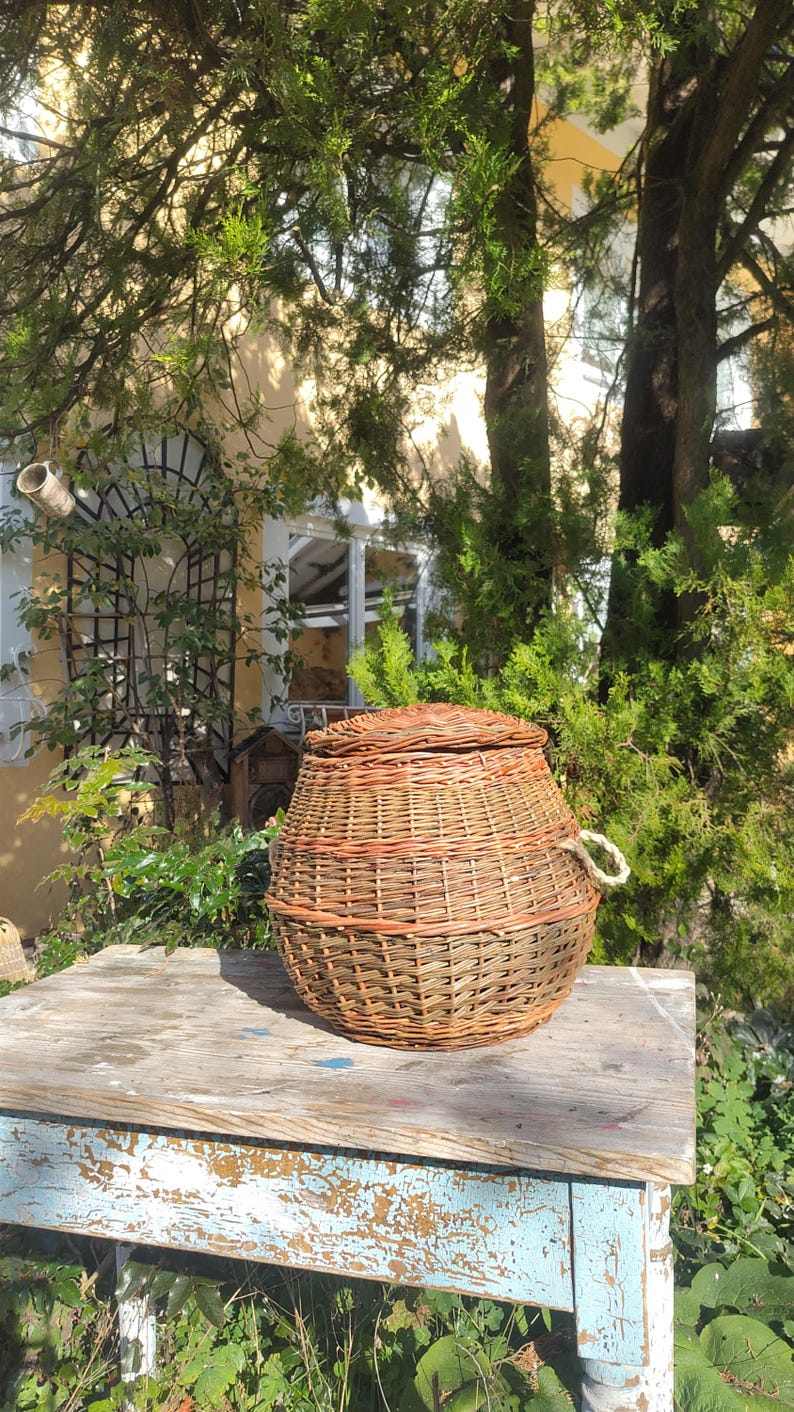 May include: A woven wicker basket with a lid and a rope handle sits on a weathered wooden table with blue paint. The basket is brown and has a natural woven pattern.