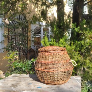 May include: A woven wicker basket with a lid and handles, sitting on a weathered wooden table with blue paint. The basket is brown and tan with a natural woven pattern.