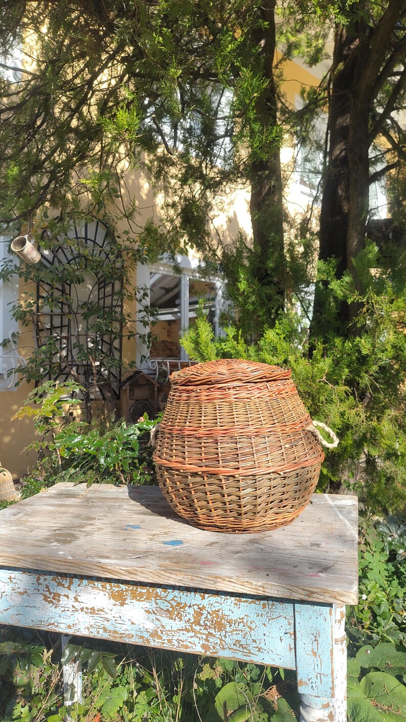 May include: A woven wicker basket with a lid and handles, sitting on a weathered wooden table with blue paint. The basket is brown and tan with a natural woven pattern.