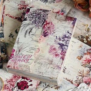 May include: A stack of rectangular paper sheets featuring a vintage floral design with purple and pink hydrangea and rose illustrations, overlaid with cursive script text. The cover sheet displays the text Flower of Memory and 160 PCS.