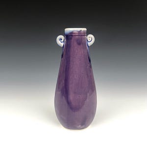 May include: A handcrafted, purple ceramic vase with a teardrop shape. The vase features two small, curled handles near the top and a white rim. The glossy finish reflects light, highlighting the rich color.