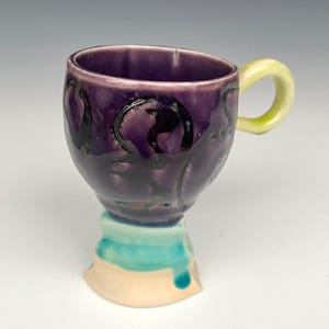 May include: A handmade ceramic mug with a purple exterior and a yellow handle. The base features turquoise and cream-colored accents. The mug has a unique, artistic design, perfect for coffee or tea.