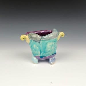 May include: A small, handmade ceramic vessel with a square shape and a vibrant color palette. The vessel features a turquoise body with a leaf-like design, purple interior, and yellow, gray, and purple accents. It has two curved handles and three small feet.
