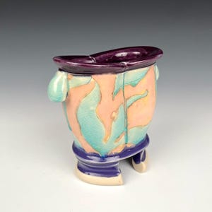 May include: A ceramic vase with a unique, abstract design. The vase features a purple rim, a peach-colored body with turquoise and yellow leaf-like patterns, and a dark purple base. Two turquoise handles and two small feet complete the design.