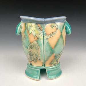 May include: A ceramic vase with a light blue rim and handles, featuring a diamond pattern in beige and teal. The vase has teal feet and is adorned with leaf-shaped carvings. The overall design is artistic and decorative.