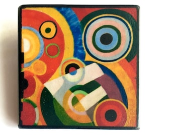 May include: A square-shaped artwork with a geometric design featuring overlapping circles in shades of red, yellow, blue, green, black, and white.