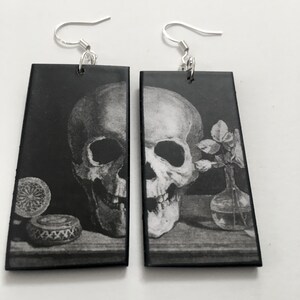 Op de afbeelding: Een paar zwarte rechthoekige oorbellen met een zilveren haak. Elke oorbel heeft een zwart-witte afbeelding van een schedel met een bloem en een fles op een tafel.