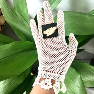 Könnte beinhalten: Weiße Häkelhandschuhe mit zarter floraler Spitzenborte. Die Handschuhe werden mit einem kleinen schwarzen quadratischen Ring mit einem Kameenbild von zwei ineinander verschlungenen Händen gezeigt.