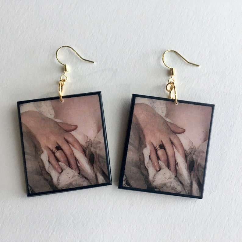 May include: A pair of square earrings with a vintage image of a hand wearing a ring. The earrings are black with a gold hook and ring.